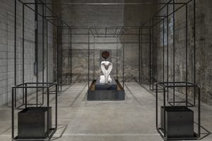 There is a hollow wire rectangular structure that takes up the whole room. The front two corners contain a metal box with liquid inside. At the center of the wire structure is a white cobra statue on a metal and dirt platform.