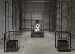 There is a hollow wire rectangular structure that takes up the whole room. The front two corners contain a metal box with liquid inside. At the center of the wire structure is a white cobra statue on a metal and dirt platform.