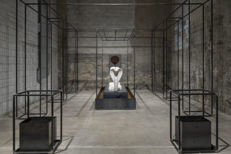 There is a hollow wire rectangular structure that takes up the whole room. The front two corners contain a metal box with liquid inside. At the center of the wire structure is a white cobra statue on a metal and dirt platform.