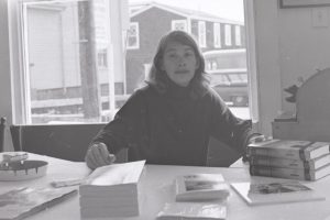 In a black and white photo, a young Mary Oliver sits at a desk surrounded by papers and books