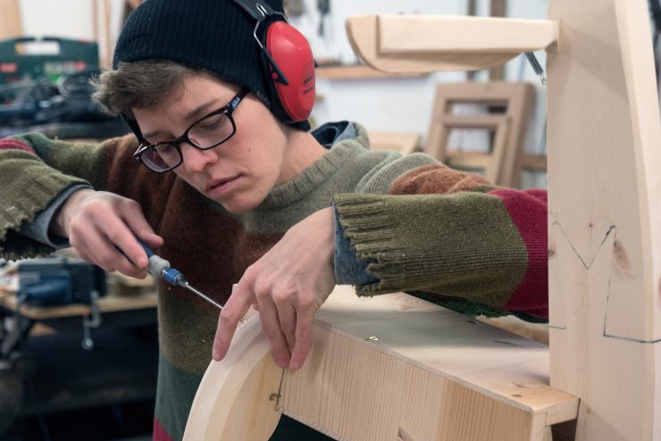 Katie is holding two pieces of wood. She is also holding a screwdriver as she joins the two pieces together.