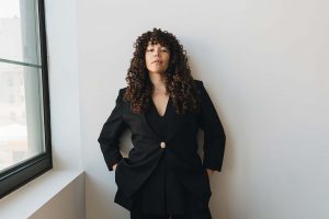 Allison stands against a wall with her hands in her pockets. Her face has a neutral expression, and she is slightly looking down at the camera.