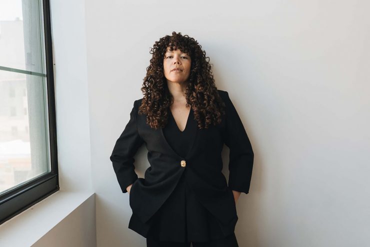 Allison stands against a wall with her hands in her pockets. Her face has a neutral expression, and she is slightly looking down at the camera.