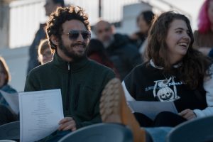 Two students, holding pieces of paper, sit in stadium seats with people around them.